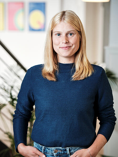 Sarah steht lächelnd vor einer Treppe in den Büroräumen von visuellverstehen. Sie trägt blondes Haar und einen blauen Pullover. 