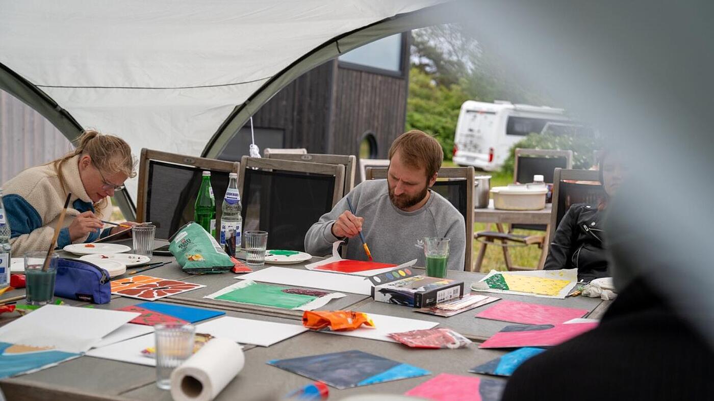 Vorm Wetter geschützt sitzen mehrere Teammitglieder von visuellverstehen unter einem Pavillon und bemalen Leinwände mit bunten Farben.