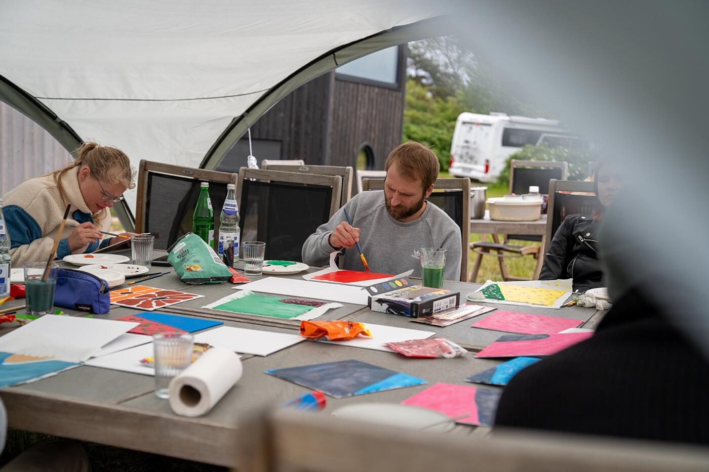 Vorm Wetter geschützt sitzen mehrere Teammitglieder von visuellverstehen unter einem Pavillon und bemalen Leinwände mit bunten Farben.