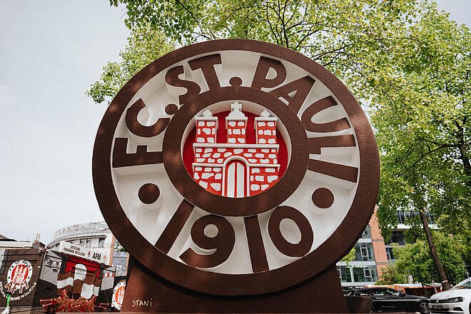 Holzskulptur des Wappens von FC St. Pauli auf einem von Bäumen umgebenen Platz, aus der Nähe fotografiert.