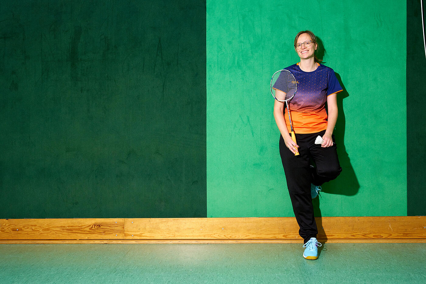 Eine Mitarbeiterin von visuellverstehen steht in Sportkleidung in einer Turnhalle und lächelt. Sie hält einen Badmintonschläger in der Hand.