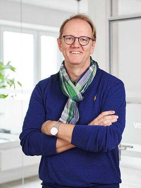 Uwe steht vor einem Besprechungsraum in den Büroräumen von visuellverstehen. Er trägt eine Brille, einen blauen Pullover und einen blau-grünen Schal.