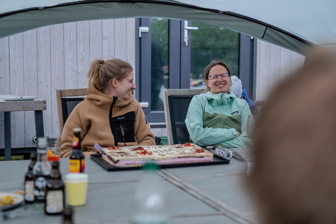 Zwei Mitarbeiterinnen von visuellverstehen sitzen unter einem Pavillon am Tisch und lachen. Vor ihnen steht ein Blechkuchen auf dem Tisch.