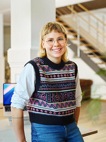 Johanna steht lächelnd vor einem Schreibtisch in den Büroräumen von visuellverstehen. Sie trägt blondes Haar, eine Brille, eine bunte Strickweste und eine blaue Jeans. 