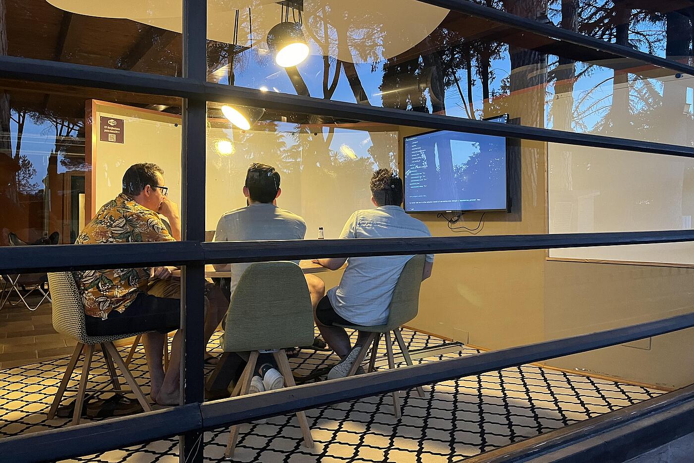 Drei Männer sitzen an einem Tisch und betrachten einen an der Wand montierten Flat Screen.