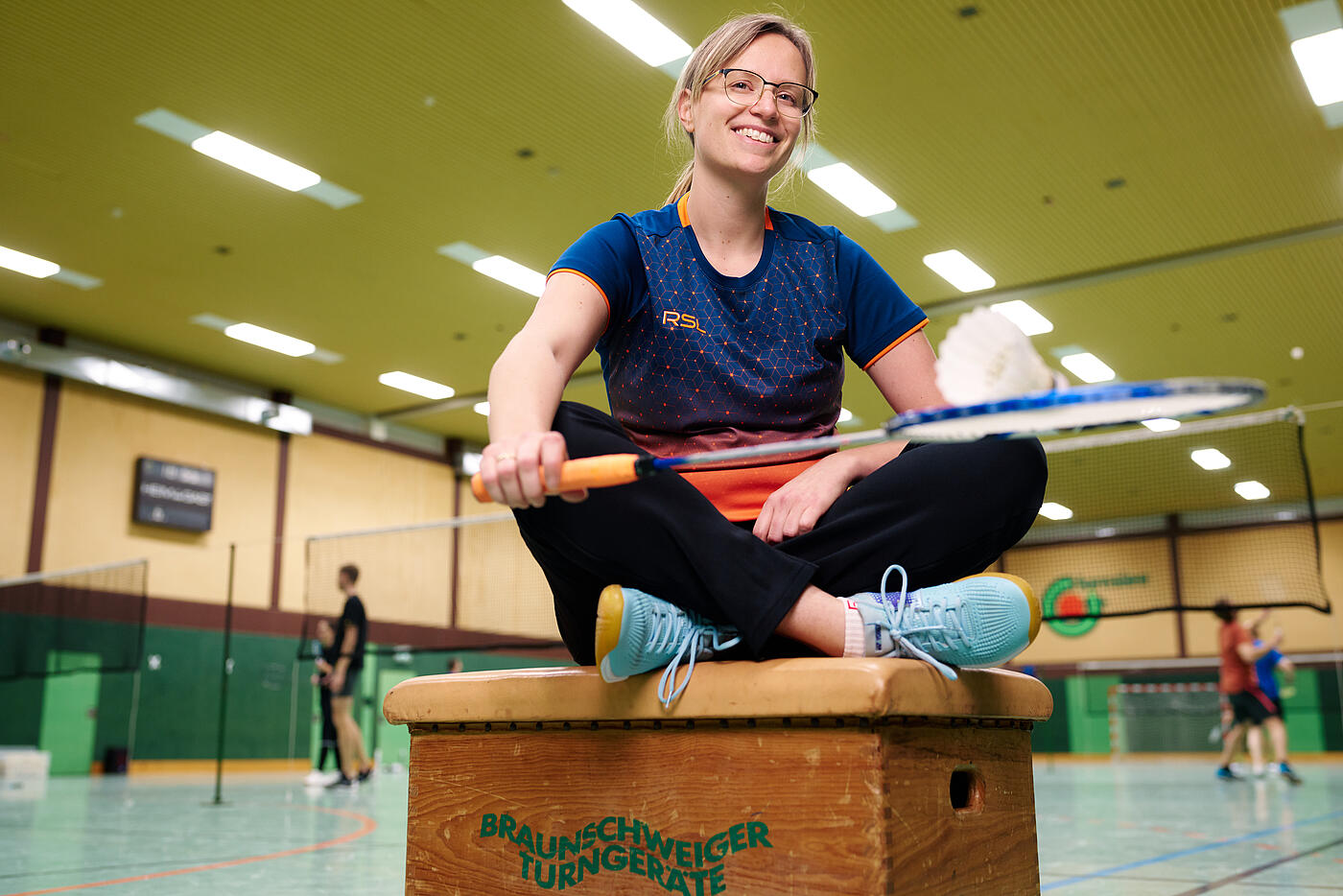 Eine Mitarbeiterin von visuellverstehen sitzt in Sportkleidung in einer Turnhalle und lächelt. Sie hält einen Badmintonschläger in der Hand.