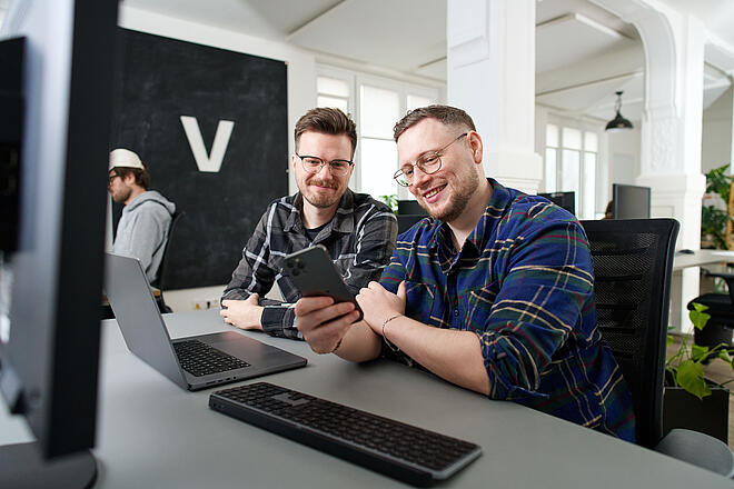 Lars und Per sitzen gemeinsam an einem Schreibtisch bei visuellverstehen und schauen auf ein Mobiltelefon.