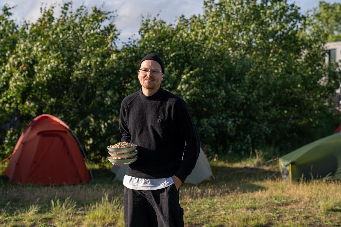 Ein Mitarbeiter von visuellverstehen steht draußen im Grünen zwischen mehreren Zelten und hat zwei Teller mit Essen in der Hand.