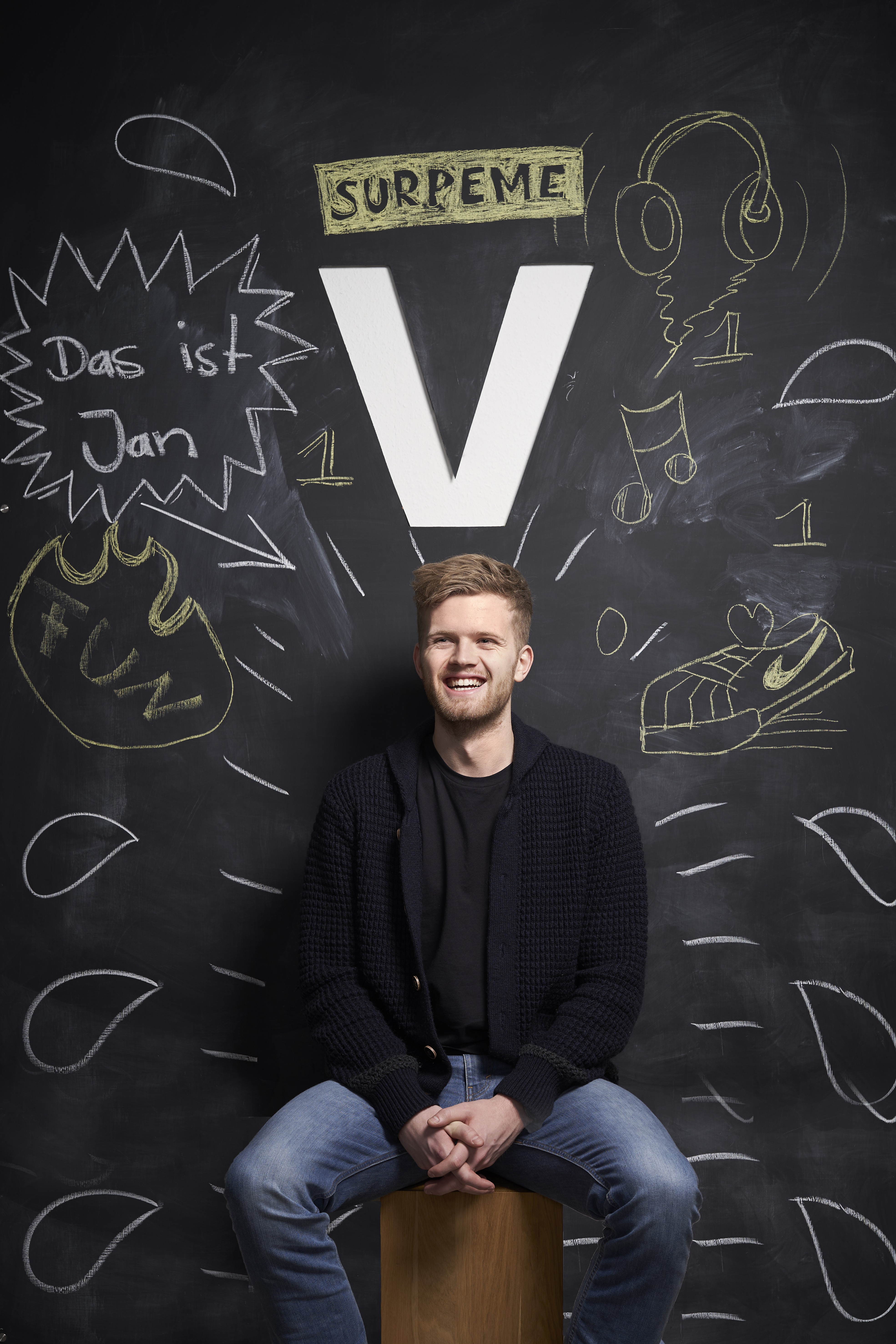 Ein Mitarbeiter von visuellverstehen sitzt lachend vor einer Tafelwand. Über seinem Kopf hängt ein großes V, und von seinem Körper gehen mit Kreide gezeichnete Linien ab.